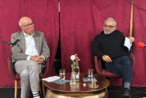 Zwei Männer auf Stühlen auf Podium, im Gespräch